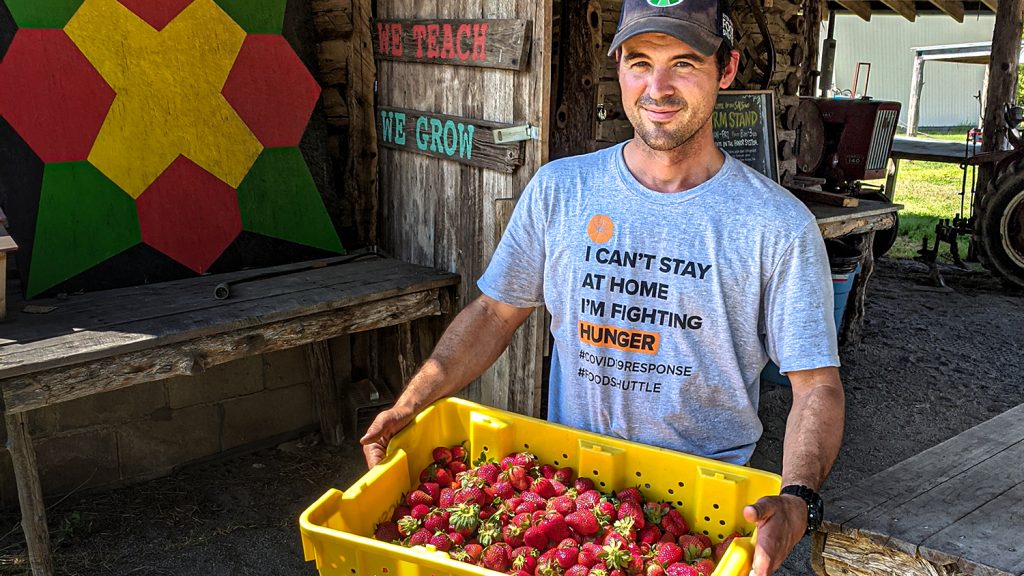 Man near a farm structure holds strawberries filling a large plastic container