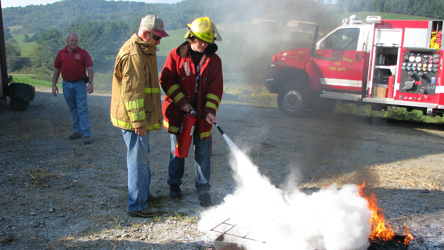 High school student uses a fire extinguisher to put out a demonstration fire with a fireman beside him and a fire truck in background.