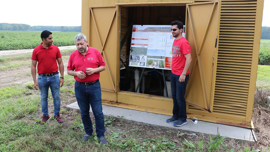 Three men in a farm field deliver a poster presentation. The poster is in a metal structure.